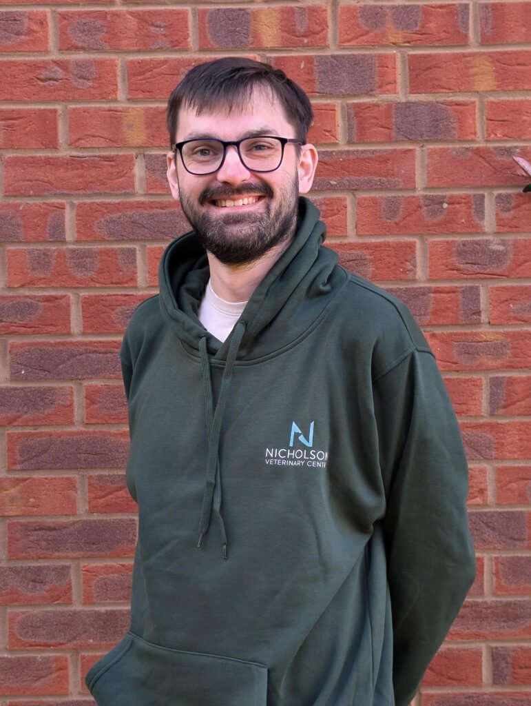A man with glasses and a beard is standing in front of a brick wall, smiling and wearing a green hoodie with "NICHOLSON VETERINARY CENTRE" written on it.