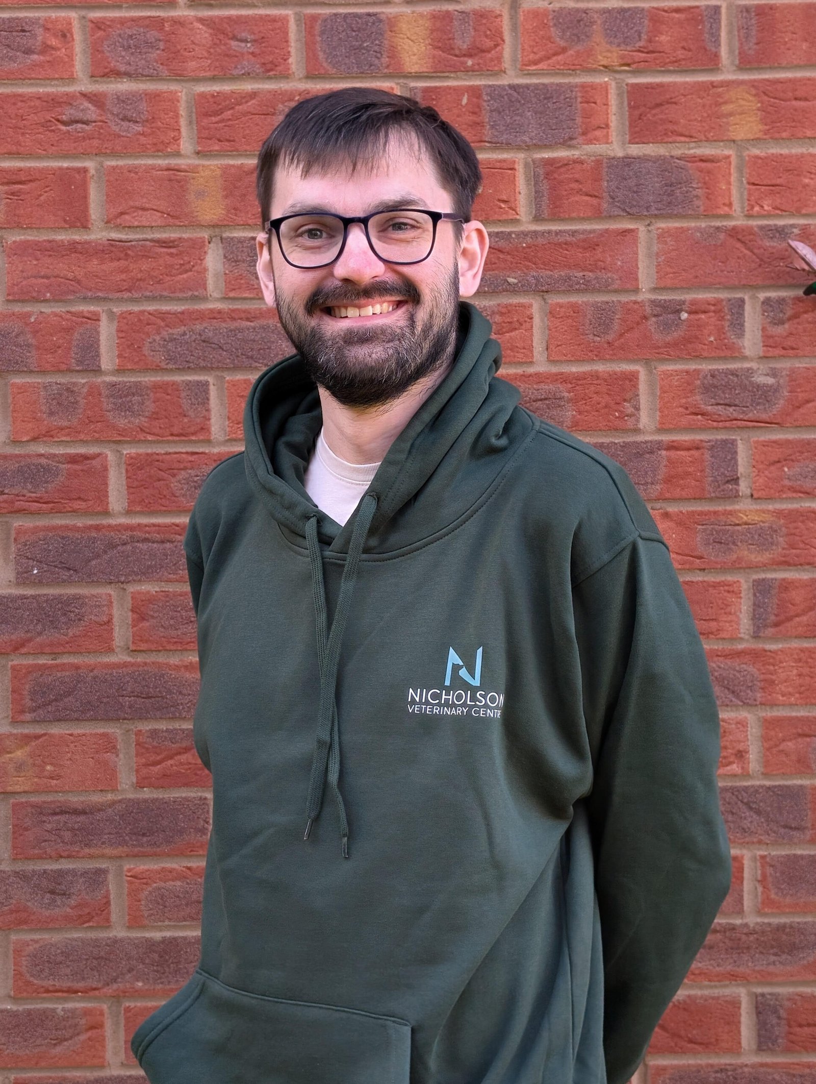 A man with glasses and a beard is standing in front of a brick wall, smiling and wearing a green hoodie with "NICHOLSON VETERINARY CENTRE" written on it.